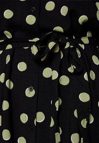 Abito nero con grandi pois verdi, dotato di una chiusura a bottoni frontale e di una cintura da annodare in vita. Il tessuto sembra leggero e testurizzato.