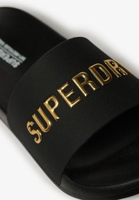 Black slide sandal featuring a textured strap with raised gold lettering spelling “SUPERDRY.” Open-toe design for easy wear.