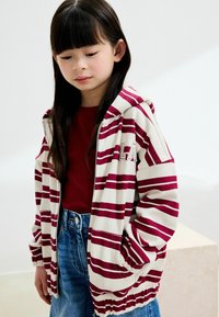 Hoodie rayé en crème et bordeaux, avec une fermeture éclair à l'avant, des poches latérales et des poignets côtelés. Associé à un top rouge et à un short en denim.