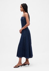 Maksi traper haljina tamnoplave boje, uski gornji dio, tanke naramenice, široka suknja s detaljima šavova, stil upotpunjen minimalističkim sandalama.