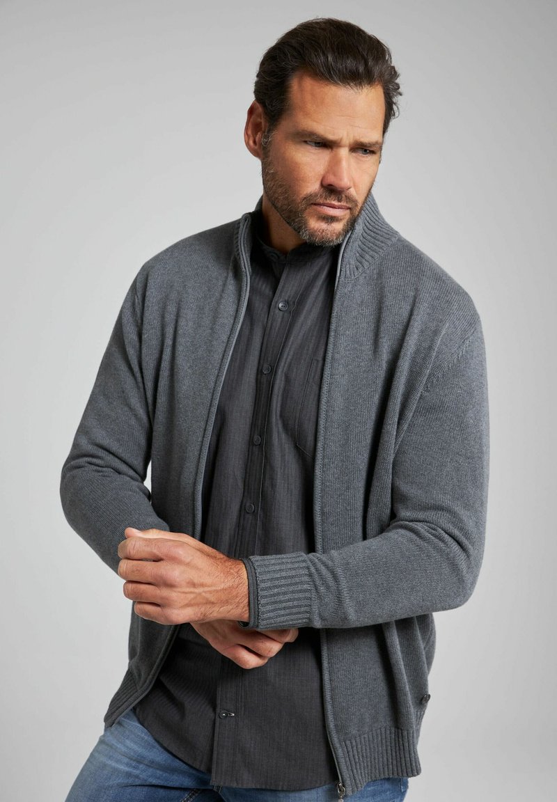 Cardigan grigio lavorato a maglia con colletto alto e polsini a coste, indossato sopra una camicia scura con bottoni. Texture liscia, design aderente.