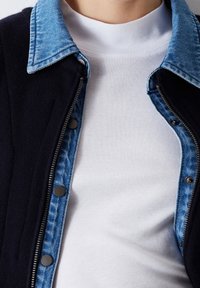 Tmavě modrá vnější bunda s džínovým límcem, vybavená stříbrným zipem a černými patentkami; vrstvená přes bílou bavlněnou košili. Strukturální tkanina.