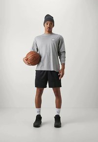 Jonge man in een grijs shirt met lange mouwen, zwarte shorts en een beanie houdt een basketbal vast, staande tegen een effen lichte achtergrond.