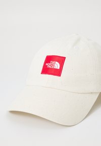 Gorra de béisbol en algodón crema con textura moteada, con un parche cuadrado rojo que dice "THE NORTH FACE" y un visera curva.