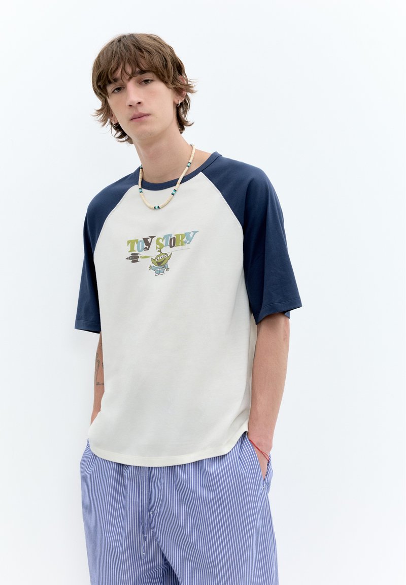 Jeune homme aux cheveux bruns en bataille portant un t-shirt Toy Story blanc et bleu marine, un pantalon bleu rayé et un collier de perles, debout les mains dans les poches.
