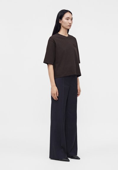 Femme aux longs cheveux noirs portant un haut ample marron foncé à manches courtes et un pantalon large bleu marine, debout devant un fond blanc uni.