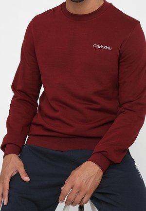 Mand iført en vinrød Calvin Klein langærmet skjorte og navy blå bukser, sidder med hænderne hvilende på lårene.