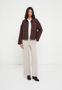 Καφέ κοντό σακάκι με μεγάλες μπροστινές τσέπες πάνω από λευκό τοπ, σετ με ανοιχτό μπεζ παντελόνι με φαρδιά μπατζάκια και μαύρες μπότες με τακούνι.