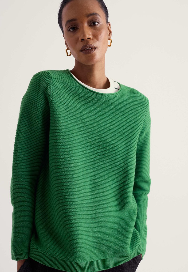 Femme aux cheveux courts portant des créoles dorées, un pull vert à côtes sur une chemise blanche, debout devant un fond uni clair.