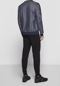 Sudadera azul marino con un diseño estampado que presenta logotipos EA7 repetidos, combinada con pantalones negros y zapatillas deportivas negras con acentos dorados.