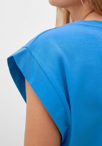 Zbliżenie na osobę w jasnoniebieskiej bluzce bez rękawów z podwiniętym mankietem, ukazującą część szyi i dużą kolczyk w kształcie obręczy.