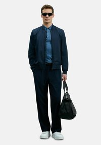 Marineblau Bomberjacke, hellblaues Poloshirt, dunkle Hose, weiße Sneakers und schwarze Tasche. Glatter Stoff, strukturierte Optik, lässige Passform.