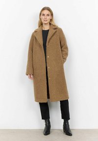 Brun, fluffig, överdimensionerad kappa med notch-krage, framknappar och sidofickor, bärs över en svart topp och svarta byxor.