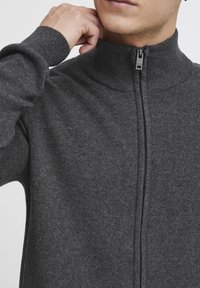 Maglione grigio scuro con zip, caratterizzato da un colletto alto e polsini a coste. Il materiale sembra morbido con una texture liscia e dettagli minimi.