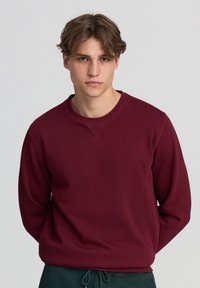 Felpa borgogna con collo rotondo, polsini e orlo a costine e una texture liscia. Il modello la indossa con pantaloni della tuta verdi.