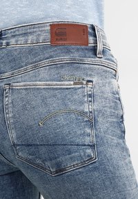 Los jeans de mezclilla azul claro presentan un parche de cuero con un logo en relieve, costuras en contraste y un diseño de arco distintivo en el bolsillo trasero.