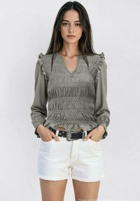 Siva bluzica s nabrusima na rukavima, V-izrezom, u kombinaciji s bijelim šortsevima. Bluzica ima teksturiranu površinu i pripijeni struk.