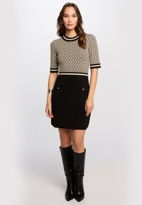 Black knit dress with short sleeves featuring a patterned top in beige and black. Dress includes two front pockets with metallic accents.