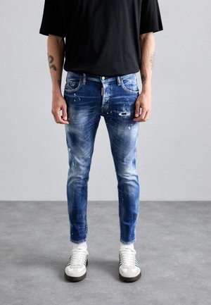 Blue denim jeans with a distressed finish, featuring paint splatters, fitted design, five pockets, and a zip fly. Paired with white sneakers.