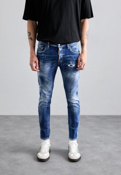 Blaue Jeans aus Denim mit einem lässigen Finish, das Farbspritzer aufweist, enganliegendem Schnitt, fünf Taschen und einem Reißverschluss. Kombiniert mit weißen Sneakern.