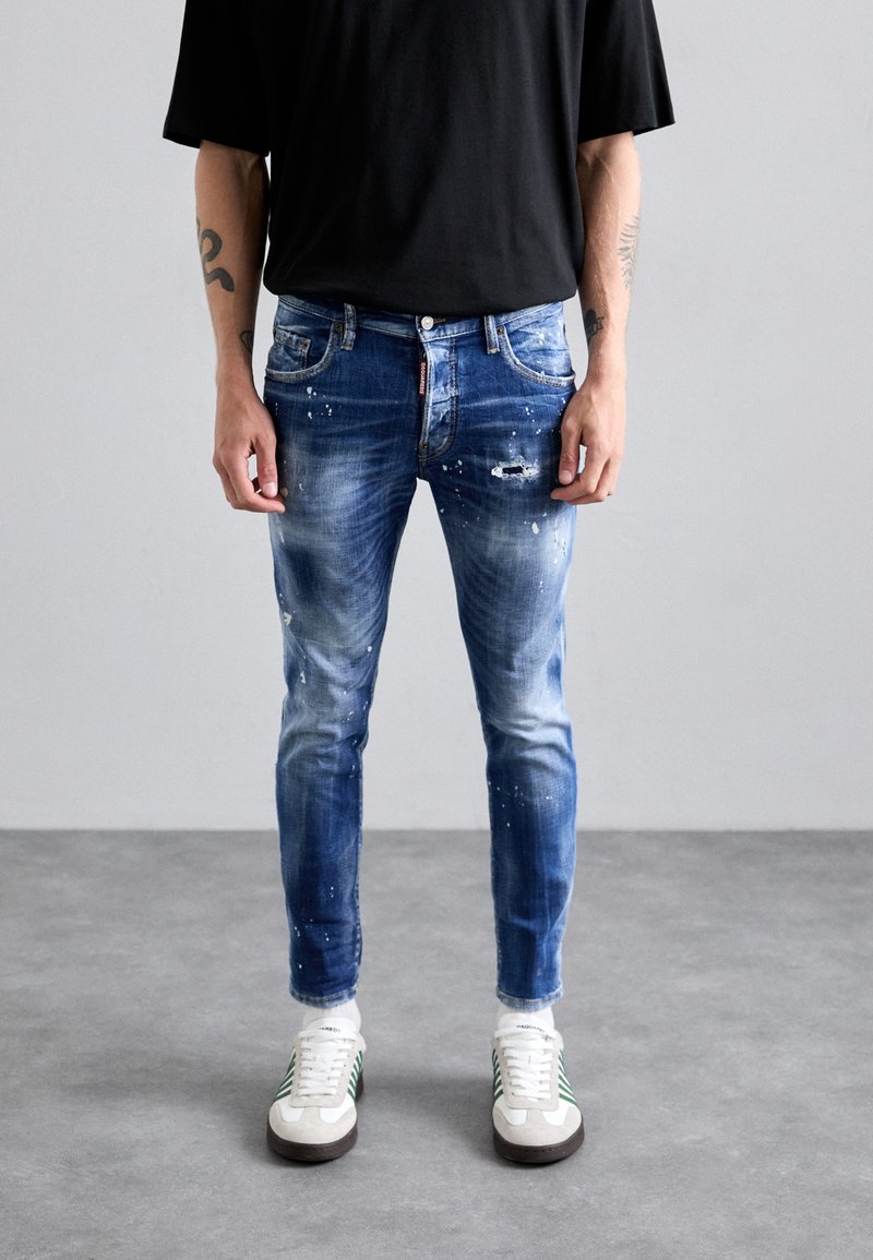 Blauwe denim jeans met een versleten afwerking, voorzien van verfspatten, een aansluitend ontwerp, vijf zakken en een ritssluiting. Gekleed met witte sneakers.