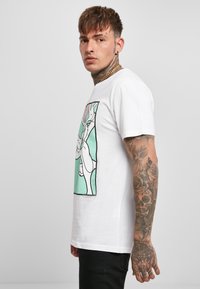 T-shirt bianca in cotone con una stampa grafica colorata sul davanti, contorno nero e scollatura a girocollo. Il modello ha dei tatuaggi sul braccio.