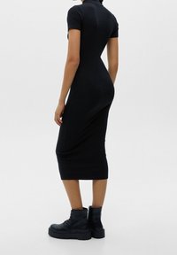 Femme portant une robe midi noire moulante à manches courtes et des bottines montantes noires épaisses à lacets, debout devant un fond uni.