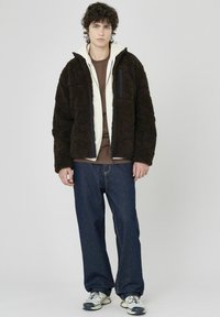 Veste en polaire marron avec un motif texturé, superposée sur un hoodie crème et une chemise marron, associée à un jean bleu foncé et des chaussures de course.