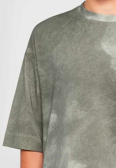 Artimas žmogaus, dėvinčio laisvai prigludusią, trumpomis rankovėmis pilką tie-dye marškinėlį su apvaliu iškirpimu, nuotrauka prieš paprastą foną.
