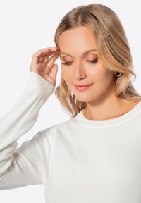 Maglione bianco a maniche lunghe con colletto e polsini a costine. Morbido al tatto, design aderente, che mostra una persona che si tocca delicatamente i capelli.
