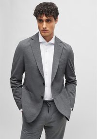 Feltett szürke textúrájú öltöny, slim fit szabással, amelynek hajtott gallérja és gombos zárása van, elöl zsebekkel, fehér inggel párosítva.