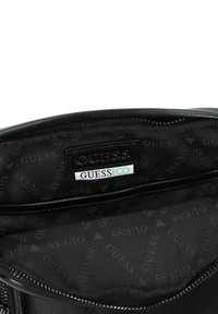Interno di borsa nera con una texture liscia, presenta un'etichetta firmata e un motivo ripetuto "GUESS" su tessuto scuro. Chiusura con zip visibile.