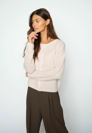Donna con lunghi capelli castani indossa un maglione rosa pallido con bottoni e pantaloni marroni, posa con una mano vicino al mento, guardando verso sinistra.