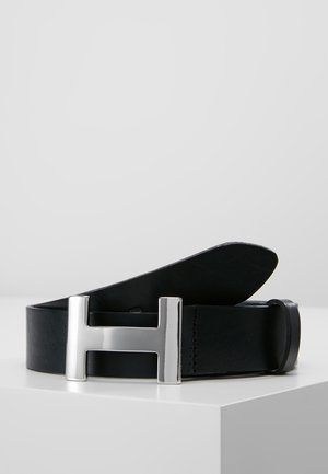 Czarny skórzany pasek z dużą srebrną metalową klamrą w kształcie litery H, pokazany zrolowany na białej powierzchni na jasnoszarym tle.