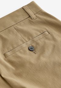 Khaki nohavice s textúrovanou tkaninou, s zadnou vreckom zabezpečeným šedým gombíkom a vyšívanými detailmi pozdĺž okrajov.