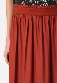 Rode rok in een rustkleur met een geplooide elastische tailleband, met een gladde textuur en een geplooid ontwerp. Dichtbijzicht van de stofdetails zichtbaar.