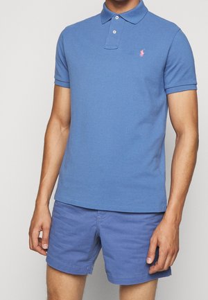 Niebieska koszulka polo o klasycznym kroju z krótkim kołnierzykiem i dwoma guzikami, z małym różowym logo na lewej piersi. Noszona z dopasowanymi szortami.