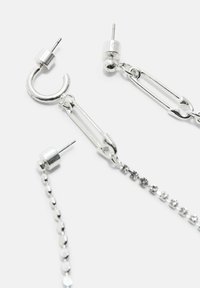 Orecchini in tonalità argento con design a spilla di sicurezza e catena su sfondo bianco, dotati di chiusura a bottone.