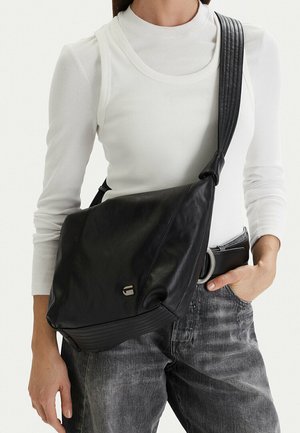 Persona vistiendo una camiseta blanca de manga larga y jeans grises, con un bolso cruzado de cuero negro que tiene una correa ancha y un pequeño logo metálico.