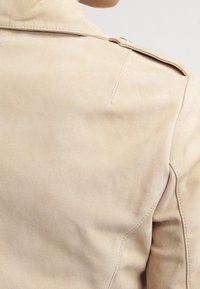 Dettaglio della spalla di una giacca in suede beige con cucitura a vista, passante e colletto indossata da una persona di spalle.