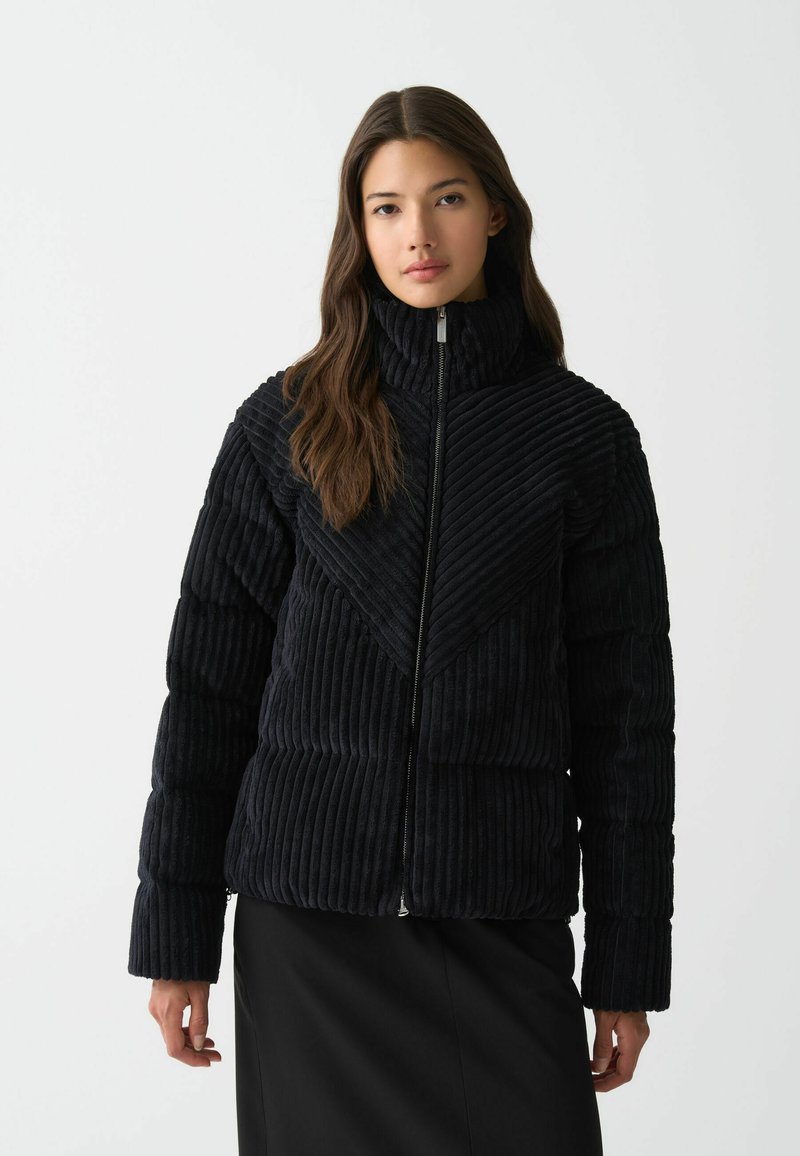 Zwarte corduroy jas met verticale ribbeltextuur, hoge kraag, ritssluiting en een geometrisch ontwerp aan de voorkant. Lange mouwen.