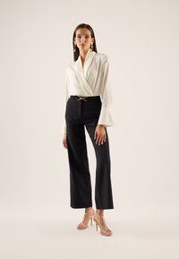 Blusa bianca avvolgente con maniche a campana, abbinata a pantaloni neri a vita alta con una cintura dorata. Sandali con tacchi a strappo dorati completano il look.