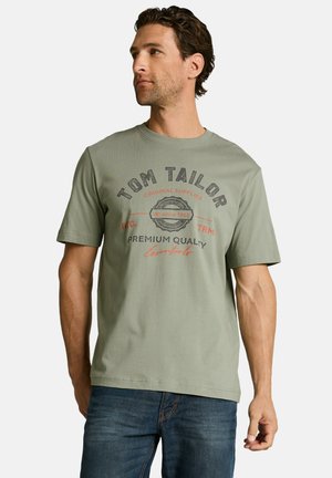 Man draagt een olijfgroen Tom Tailor T-shirt met bedrukt tekst en blauwe spijkerbroek, kijkt naar links tegen een witte achtergrond.