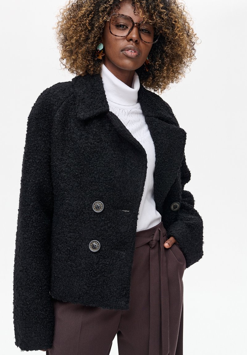 Czarny, teksturowany płaszcz dwurzędowy z dużym kołnierzem i dwoma guzikami; noszony na białym golfie, w połączeniu z brązowymi spodniami z wysokim stanem.