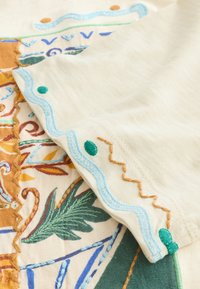 Närbild av vikt tyg med broderade botaniska och geometriska mönster i blå, gröna, bruna och orange nyanser på krämfärgad bakgrund.