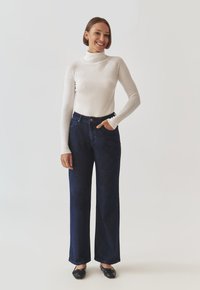 Maglione bianco a collo alto, jeans larghi blu scuro e scarpe nere basse. La modella posa con una mano in tasca, mostrando un outfit casual.