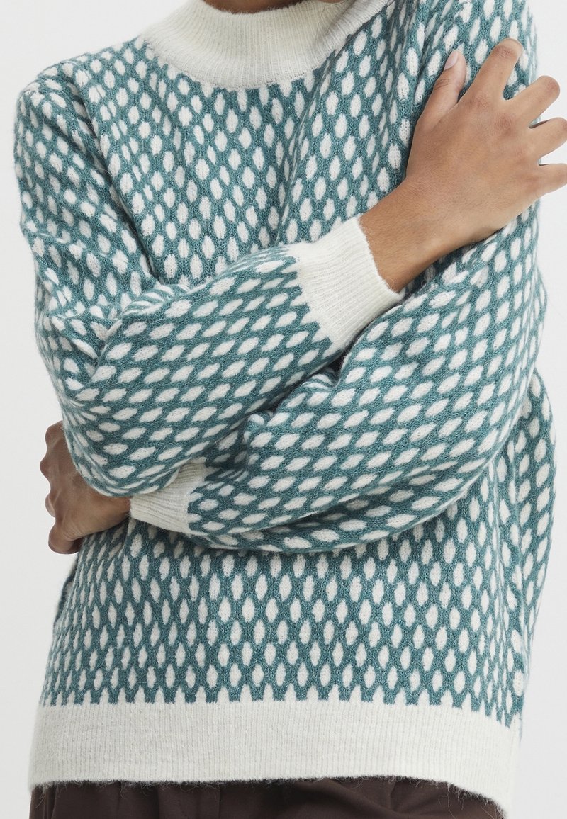 Pull-over en maille présentant un motif losange turquoise et blanc, avec un col montant et des poignets et un ourlet côtelés contrastants en crème.