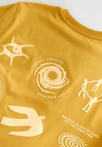 Tissu jaune moutarde avec des motifs abstraits crème : spirale avec « The Next Dimension », figures stylisées, forme d’oiseau, et texte « Believe in everything at the same time. »