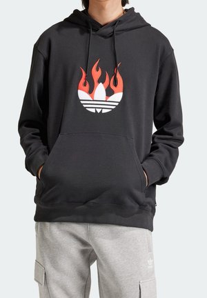 Osoba ubrana w czarną bluzę z kapturem z płonącym logo Adidas na piersi oraz jasnoszare spodnie dresowe Adidas z kieszeniami.