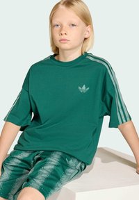 Zelené voľné tričko s krátkym rukávom, s malým logom na hrudi a vzorované zelené šortky so bočnými pruhmi.